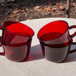 May include: Four ruby red glass coffee cups are stacked in pairs. Each cup has a rounded handle and a slightly angled rim. The cups are sitting on a light-colored surface, with a blurred outdoor background.