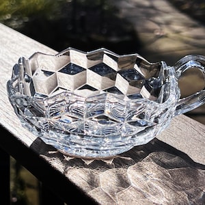 May include: A clear, faceted glass teacup with a handle. The cup has a geometric pattern and sits on a wooden surface, with sunlight casting shadows. The cup is empty and appears to be vintage.