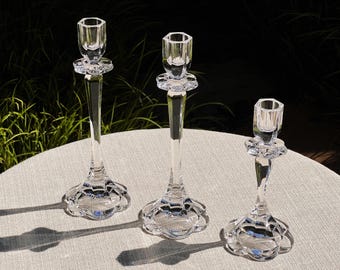 May include: Three clear glass candle holders of varying heights with a decorative, scalloped base. The candle holders are arranged on a light gray surface.
