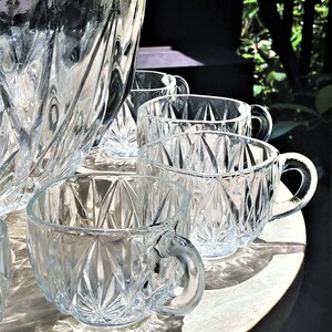 May include: A clear glass punch bowl and matching cups with a diamond pattern. The punch bowl has a scalloped rim. Several cups are arranged on a round, light-colored wooden tray. The cups have handles.