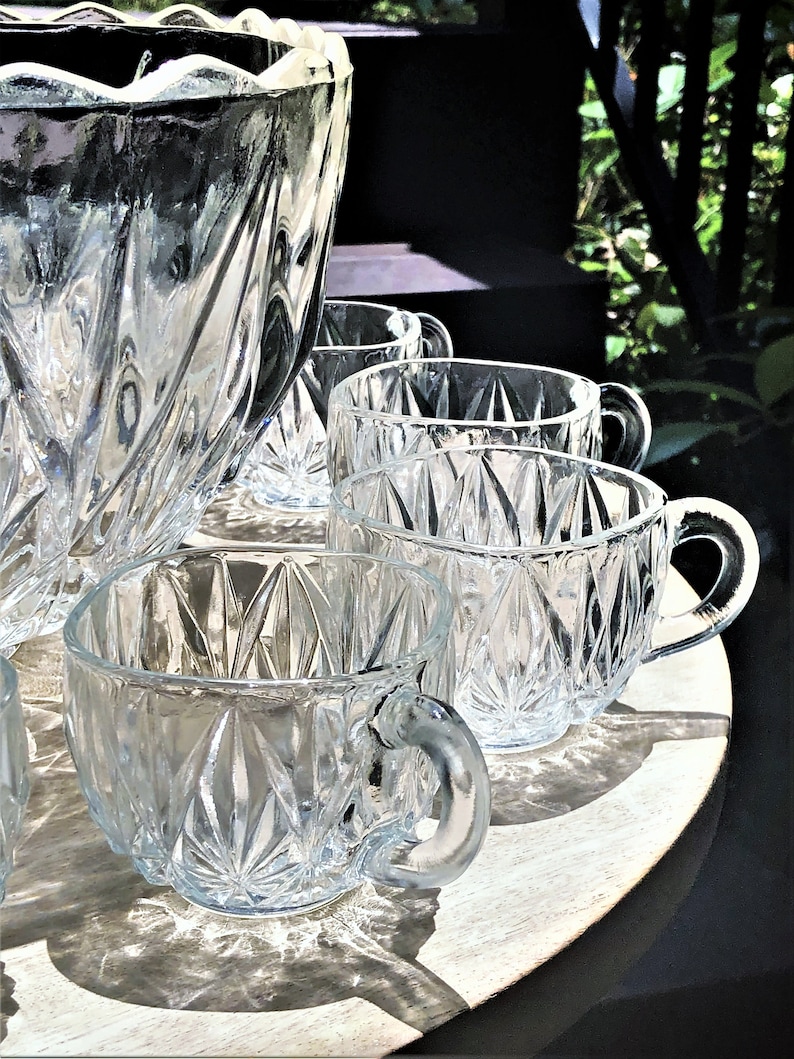May include: A clear glass punch bowl and matching cups with a diamond pattern. The punch bowl has a scalloped rim. Several cups are arranged on a round, light-colored wooden tray. The cups have handles.