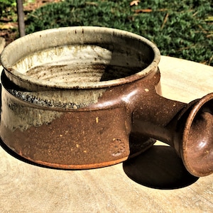 May include: A brown and white ceramic bowl with a single handle. The bowl has a speckled glaze and a slightly rounded shape. The handle is attached to the side of the bowl and has a flared opening.
