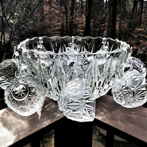 May include: A clear, cut-glass punch bowl set with a large bowl and several matching cups. The bowl and cups have a geometric starburst pattern. The set is displayed on a wooden surface with a blurred forest background.
