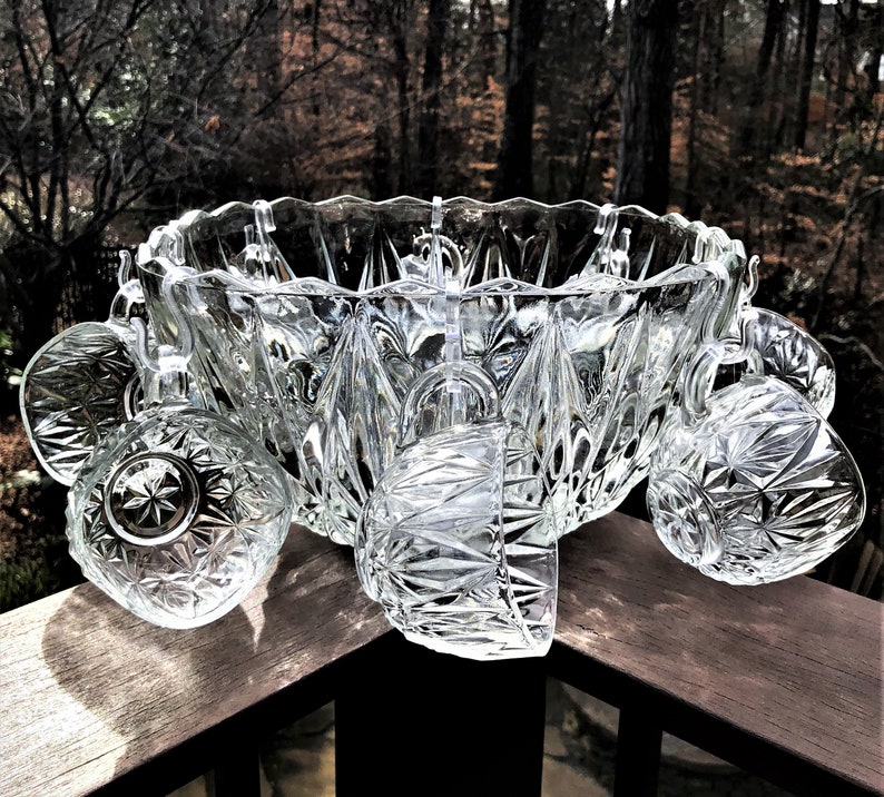 May include: A clear, cut-glass punch bowl set with a large bowl and several matching cups. The bowl and cups have a geometric starburst pattern. The set is displayed on a wooden surface with a blurred forest background.