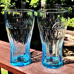 May include: Two clear Coca-Cola glasses with a light blue tint. The glasses have a classic shape with the Coca-Cola logo embossed on the front. They are on a wooden surface, with a blurred outdoor background.