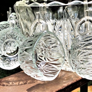 May include: Clear glass punch bowl set with matching cups. The glass has a starburst pattern and is displayed on a wooden surface. The set includes a large bowl and several handled cups, perfect for serving beverages.