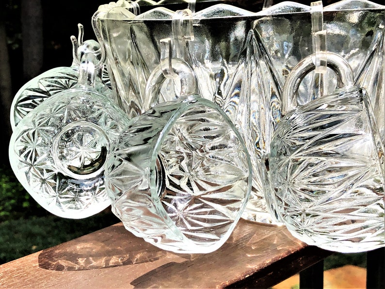 May include: Clear glass punch bowl set with matching cups. The glass has a starburst pattern and is displayed on a wooden surface. The set includes a large bowl and several handled cups, perfect for serving beverages.