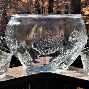 May include: A clear glass punch bowl with a raised rose design. The bowl is surrounded by four matching glass cups.