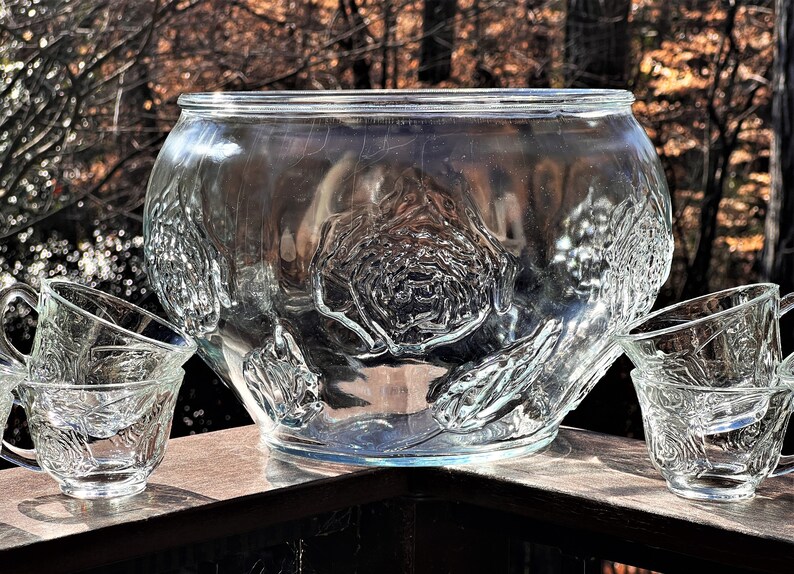 May include: A clear glass punch bowl with a raised rose design. The bowl is surrounded by four matching glass cups.