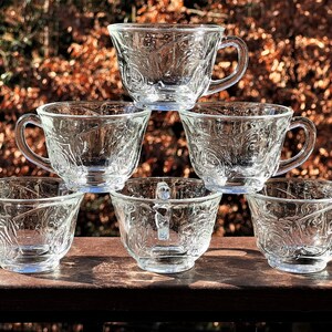 May include: Six clear glass teacups with a delicate floral pattern stacked in a pyramid shape. The cups have a rounded shape and a handle on the side.