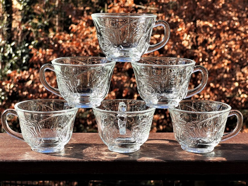 May include: Six clear glass teacups with a delicate floral pattern stacked in a pyramid shape. The cups have a rounded shape and a handle on the side.