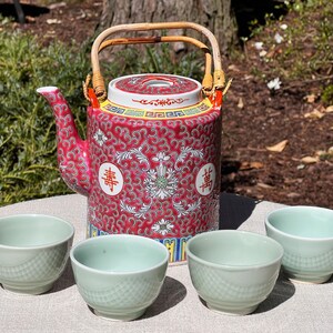 May include: A red and white patterned teapot with a bamboo handle and four light green teacups. The teapot has Chinese characters and a decorative design. The teacups are small and have a textured surface.