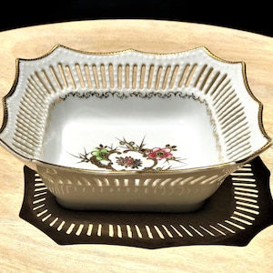 May include: A white porcelain bowl with a square shape and a delicate openwork design. The bowl has a gold rim and a floral pattern in shades of pink, green, and gold.