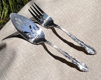 May include: Silver serving fork and pie server with ornate handles. The pie server has a decorative cut-out design. Both utensils are placed on a neutral-colored linen surface, with shadows cast by sunlight.