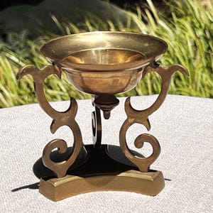 May include: A polished brass bowl with a smooth, rounded interior and a wide rim, supported by a decorative stand featuring three ornate, curved legs that spiral at the base and connect to a central, triangular, tiered pedestal structure.