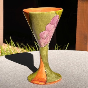 May include: A green and orange ceramic goblet with a painted design of purple grapes. The goblet has a wide, flared bowl and a tall, slender stem.