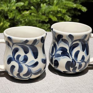 May include: Two ceramic mugs with a rounded, bulbous shape and a glossy cream-colored finish. Each mug features a hand-painted pattern of swirling, organic leaf-like shapes in varying shades of dark navy blue and soft light blue across the exterior.