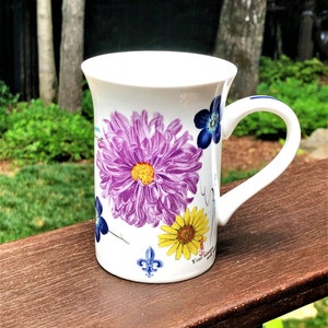 May include: White ceramic mug with a floral design featuring purple, yellow, and blue flowers. The mug has a handle and the text "Your garden will always be with you" on the bottom.