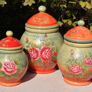 May include: Set of three ceramic canisters with lids, featuring a floral design with roses in shades of pink and red. The canisters have a green and orange color scheme with a distressed finish.