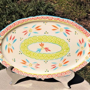 May include: A white oval serving platter with a scalloped edge. The platter is decorated with a colorful floral pattern in shades of red, orange, yellow, green, blue, and white. The platter is on a silver stand.