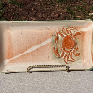 May include: Rectangular ceramic serving dish with a crab design. The dish has a light peach and white speckled background with a white diagonal stripe. The crab is orange. The rim is light green.