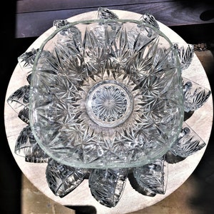 May include: A clear glass punch bowl set with a square bowl and starburst pattern. The bowl is surrounded by matching cups, all resting on a light-colored wooden surface. The set is designed for serving beverages.