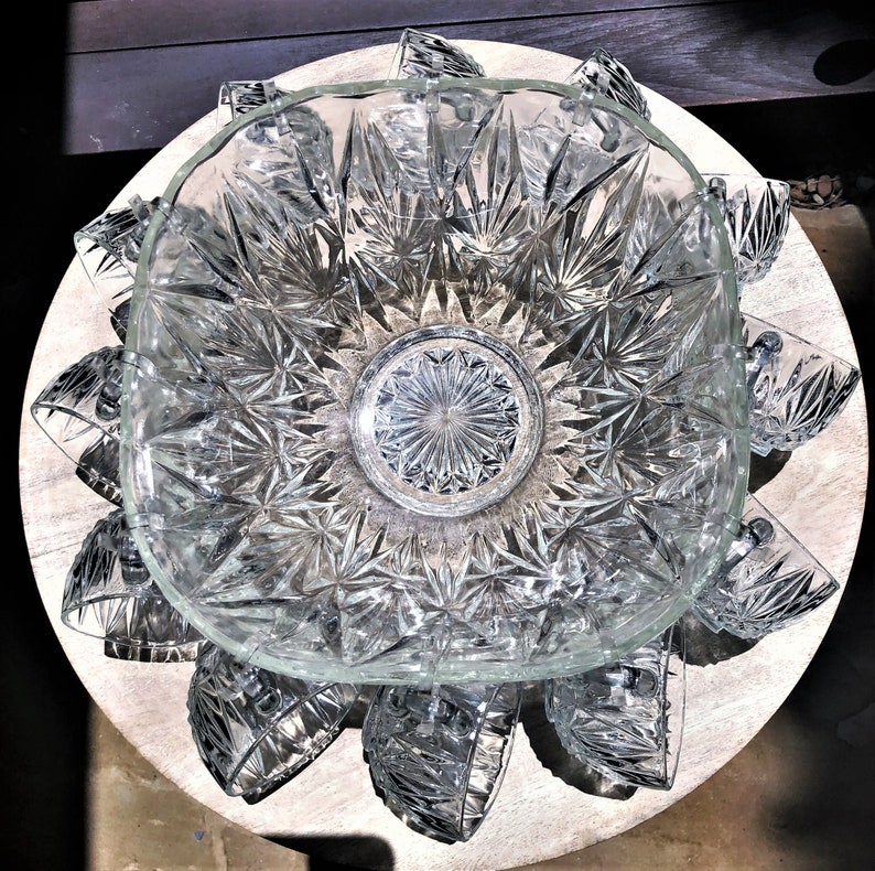 May include: A clear glass punch bowl set with a square bowl and starburst pattern. The bowl is surrounded by matching cups, all resting on a light-colored wooden surface. The set is designed for serving beverages.