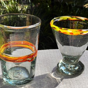 May include: Two clear glass drinking glasses with orange and yellow painted bands around the top rim. The glass on the left is a tall, cylindrical glass. The glass on the right is a shorter, wider glass with a flared base.