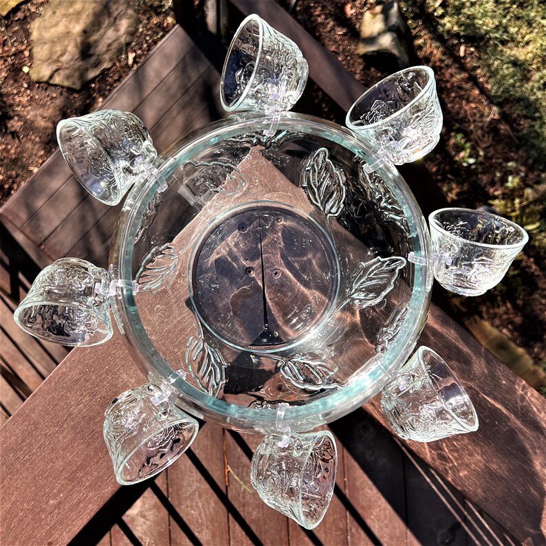 May include: A clear glass punch bowl with a leaf design and eight small glass cups attached around the rim. The bowl is filled with water and the cups are empty.