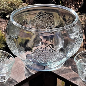 May include: A clear glass punch bowl with a rose pattern and two matching cups. The bowl is large and has a wide rim. The cups are smaller and have a handle.