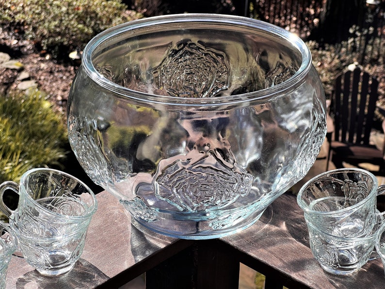 May include: A clear glass punch bowl with a rose pattern and two matching cups. The bowl is large and has a wide rim. The cups are smaller and have a handle.