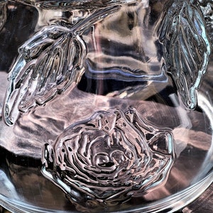May include: A clear glass bowl with a textured design of roses and leaves. The bowl is empty and has a reflective surface.