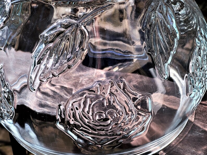 May include: A clear glass bowl with a textured design of roses and leaves. The bowl is empty and has a reflective surface.