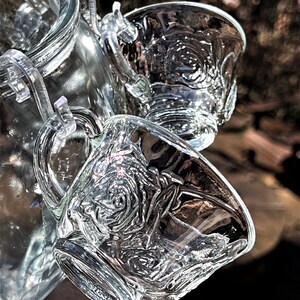 May include: A set of clear glass cups with a rose pattern. The cups are attached to a glass pitcher with a clear plastic lid.