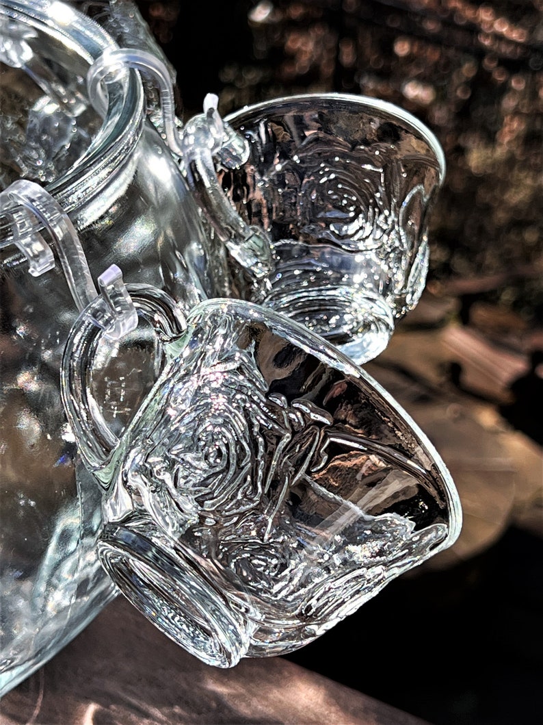 May include: A set of clear glass cups with a rose pattern. The cups are attached to a glass pitcher with a clear plastic lid.