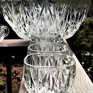 May include: A clear glass punch bowl set with a large bowl and several small cups. The bowl and cups have a diamond-cut pattern. The set is displayed on a wooden surface, with a blurred background of greenery.