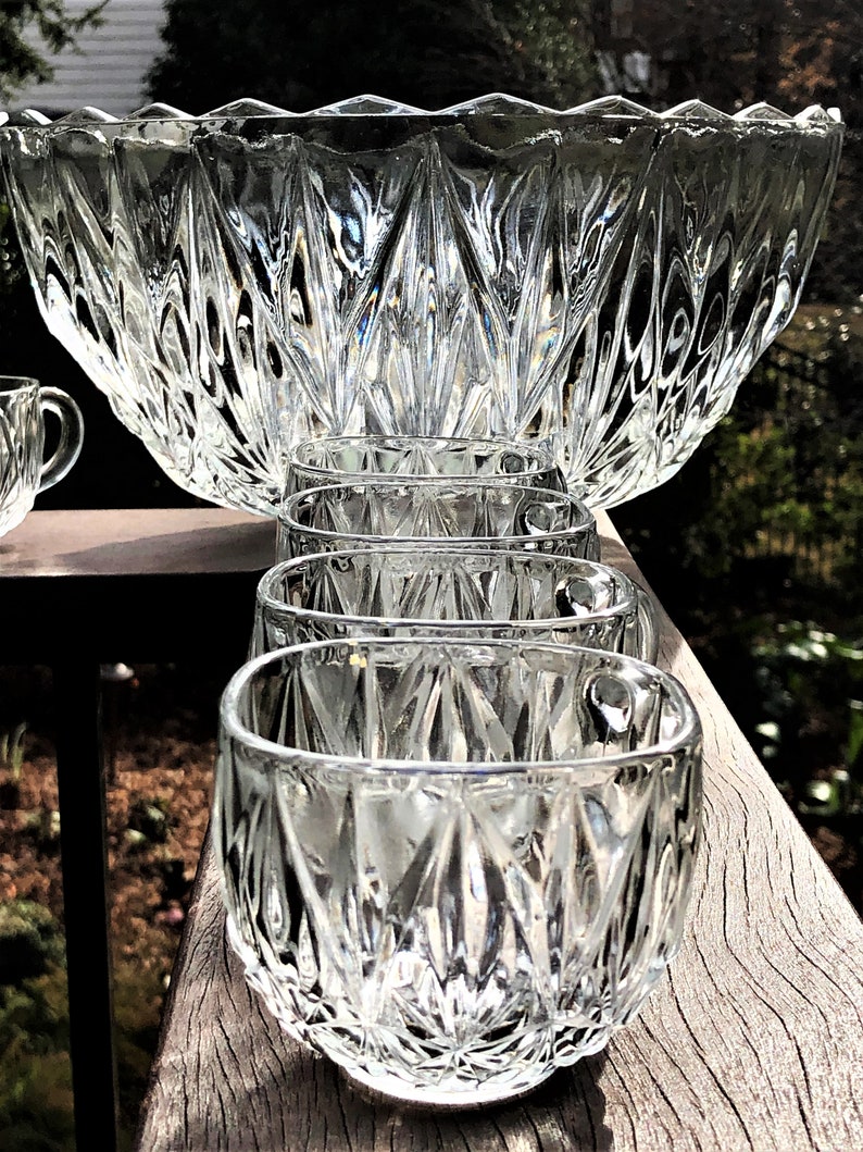 May include: A clear glass punch bowl set with a large bowl and several small cups. The bowl and cups have a diamond-cut pattern. The set is displayed on a wooden surface, with a blurred background of greenery.