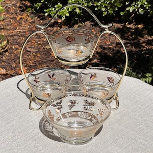 May include: A vintage serving set with four clear glass bowls, each adorned with gold and burgundy leaf and floral designs. The bowls are held in a gold-toned metal stand with a handle, perfect for serving snacks or desserts.