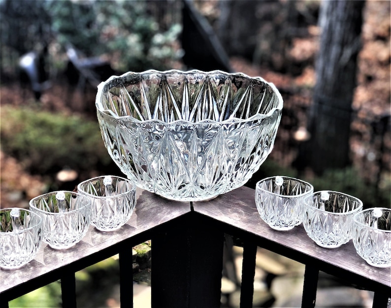 May include: A clear glass punch bowl set with a large bowl and six small square-shaped glasses. The bowl and glasses have a diamond-cut pattern. The set is displayed on a wooden surface with a blurred outdoor background.