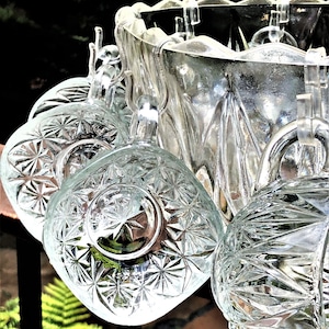 May include: A clear glass punch bowl set with six matching cups. The cups and bowl have a starburst pattern. The cups hang from the rim of the bowl. The background is blurred with green foliage.