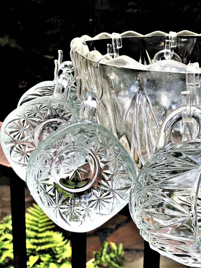 May include: A clear glass punch bowl set with six matching cups. The cups and bowl have a starburst pattern. The cups hang from the rim of the bowl. The background is blurred with green foliage.