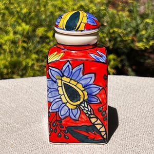May include: A rectangular ceramic jar with a lid. The jar is painted red with a large blue and yellow floral design. The lid is yellow, blue, and green. The jar is sitting on a light gray surface with a green and brown background.