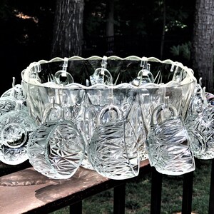 May include: A clear glass punch bowl set with a scalloped rim, holding several clear glass cups. The cups are hanging from the bowl's rim. The set is displayed on a wooden surface, with a natural background.