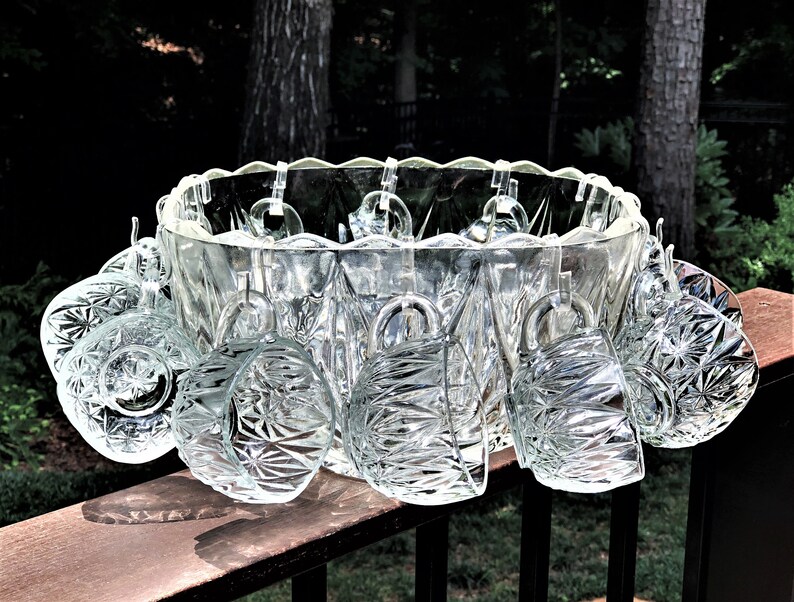 May include: A clear glass punch bowl set with a scalloped rim, holding several clear glass cups. The cups are hanging from the bowl's rim. The set is displayed on a wooden surface, with a natural background.