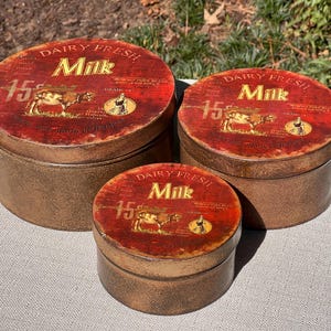 May include: Three vintage-style round metal containers with lids. The lids are red with gold lettering that reads "Dairy Fresh Milk" and feature a cow illustration. The containers are various sizes and have a distressed, aged appearance.