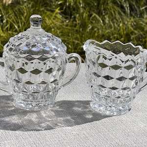 May include: A clear, vintage sugar bowl with a lid and a creamer. Both pieces are made of glass and feature a diamond-cut pattern. The sugar bowl has two handles, and the creamer has a scalloped rim and a handle. The set is on a light-colored surface.