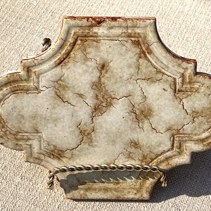 May include: A decorative ceramic tile with a white background and brown marbling. The tile has a scalloped edge and is displayed on a gold stand.