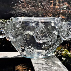 May include: A clear glass punch bowl with a decorative design and six matching cups hanging from the rim. The bowl and cups are made of clear glass and have a textured surface.