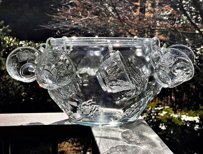 May include: A clear glass punch bowl with a decorative design and six matching cups hanging from the rim. The bowl and cups are made of clear glass and have a textured surface.