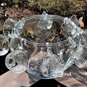 May include: A clear glass punch bowl with a set of matching cups hanging from the rim. The cups have a textured design and are perfect for serving punch at a party.